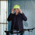 Een man die zijn gele fietshelm rechtzet terwijl hij voor een fiets staat, met op de achtergrond een blauwe muur.